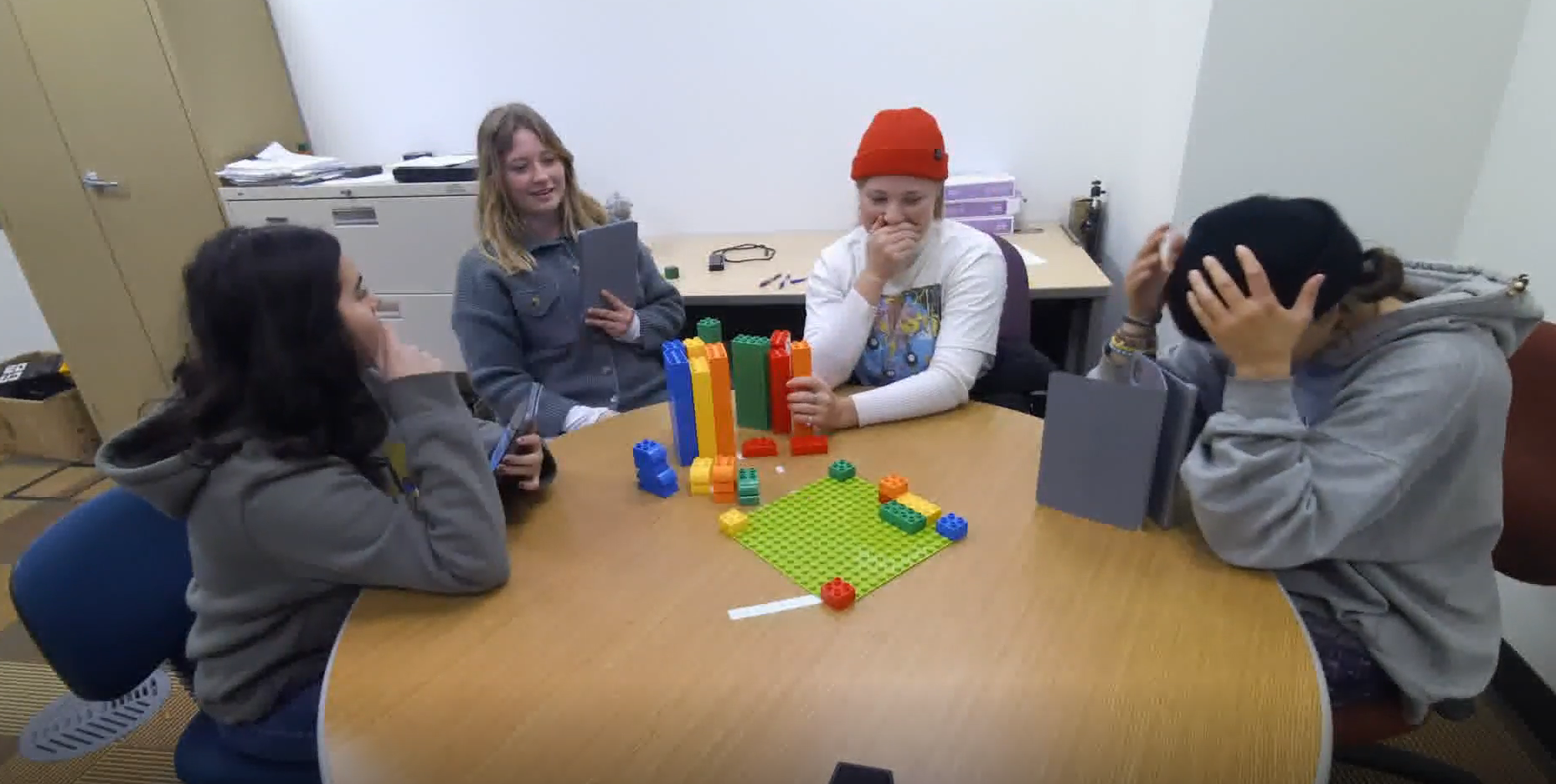 Image of four participants completing the DPIP task, along with relevant dialogue, where participants disagree about whether an orange block is next. One participant rests her head on her hands.