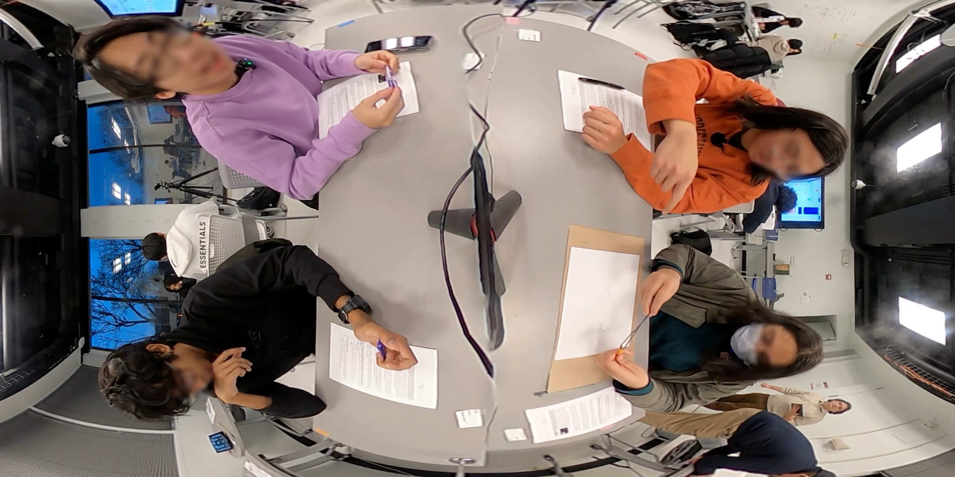 Top-down classroom frame with faces obscured, showing a student making a hand gesture near the table.