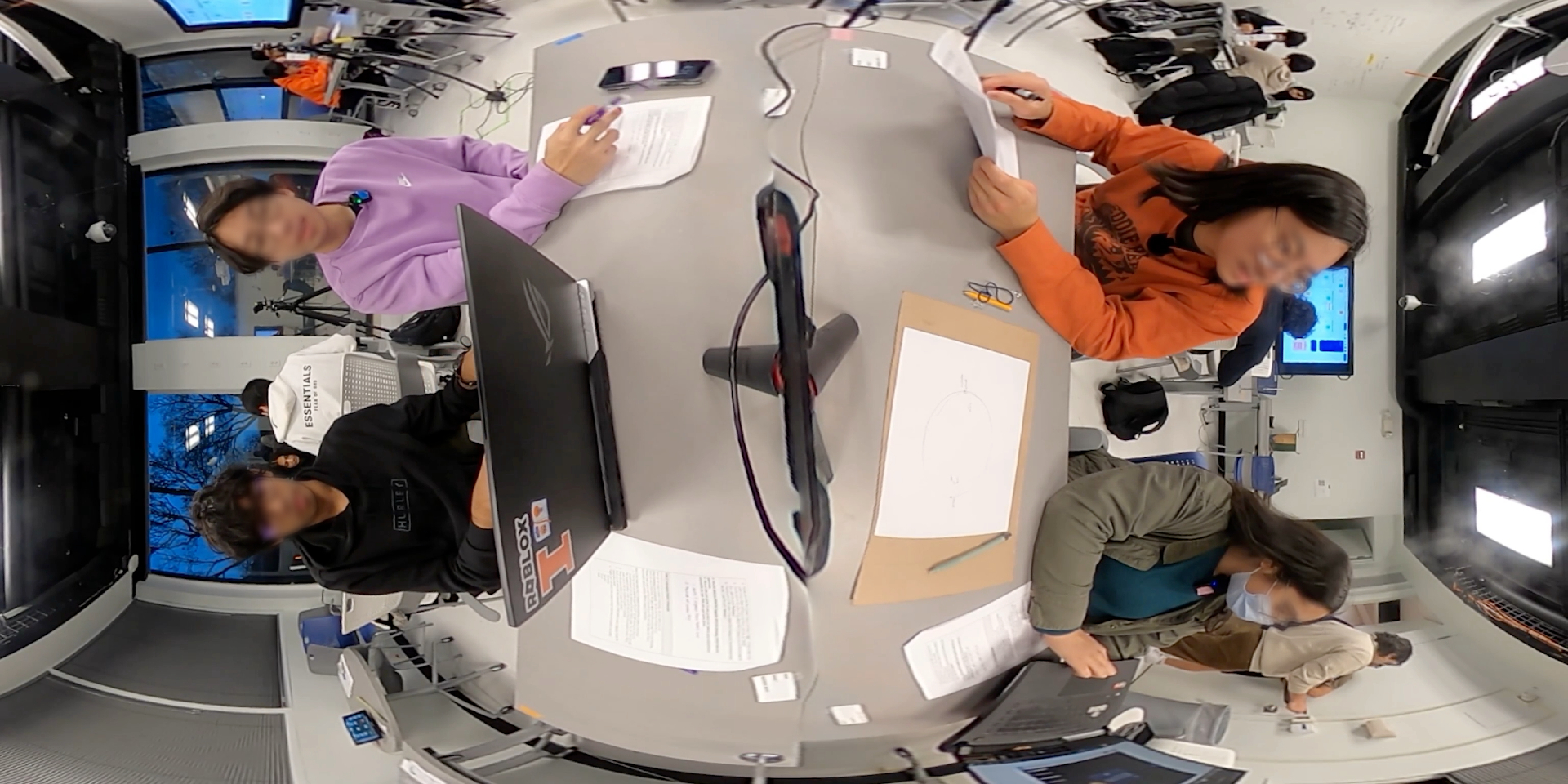 Top-down classroom frame with faces obscured, showing students arranged around shared laptops and worksheets.